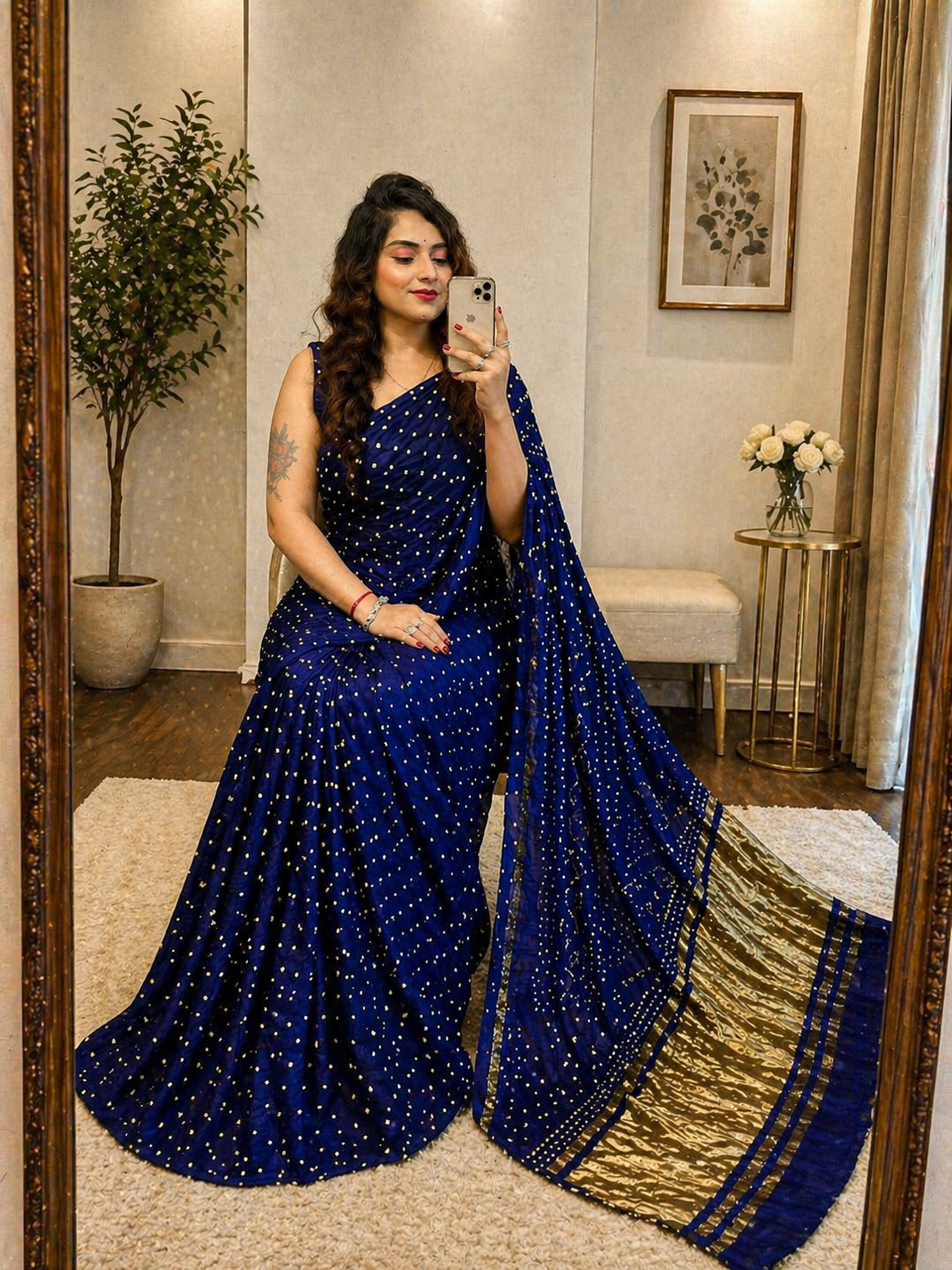 Woman in a blue saree with gold border taking a mirror selfie in a home setting.